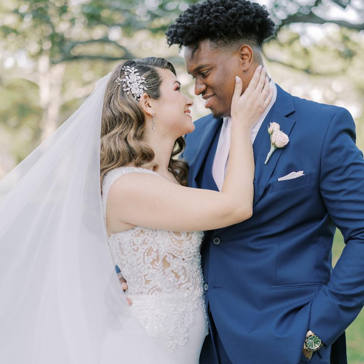 image of a bride in a white lace dress with a long veil and groom wearing a blue suit with a cream rose in his lapel staring into each others eyes and smiling