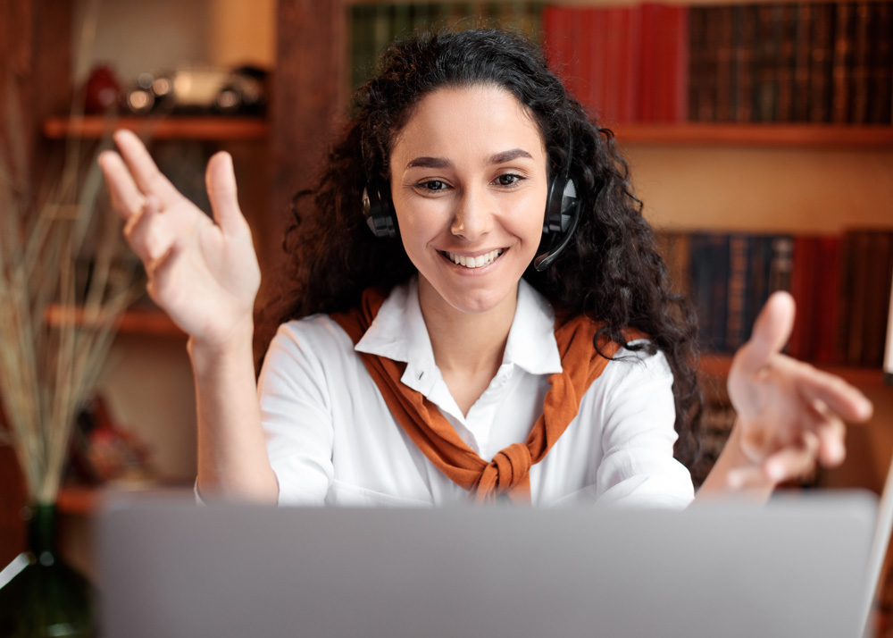 A smiling young woman with long dark brown curly hair talks to someone on a video call while gesturing with her hands.